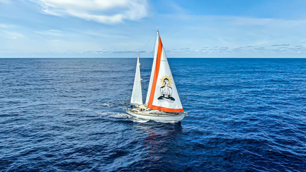 SV Delos sailing through the open sea. The sails are up, the water is calm, and the sky is blue