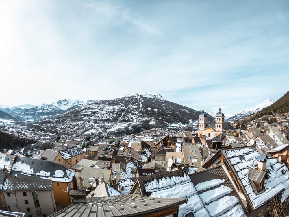 View of Briançon old town from on top of the old barracks. snow capped mountains sit in the background. the sky is blue. the roofs of the town are covered in snow.