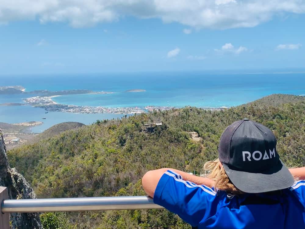 enfant avec une casquette à l'envers sur laquelle est écrit "roam" regardant un magnifique paysage de collines boisées vertes et d'eau bleu électrique