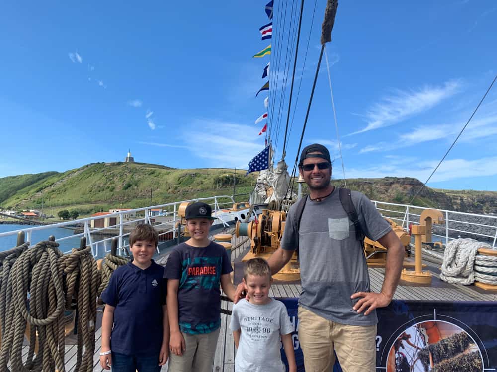 three kids and dad standing on deck of a sailing boat moored near the shore. The sky is bright blue and the weather is fabulous. They're all smiling and have their arms around each other.