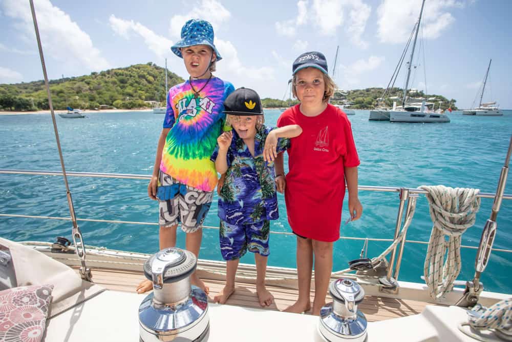 three kids decked out in colourful shirts, standing on the deck of their sail boat. The boat is floating on incredible turquoise water. There are a few other sailing vessels floating i the background.