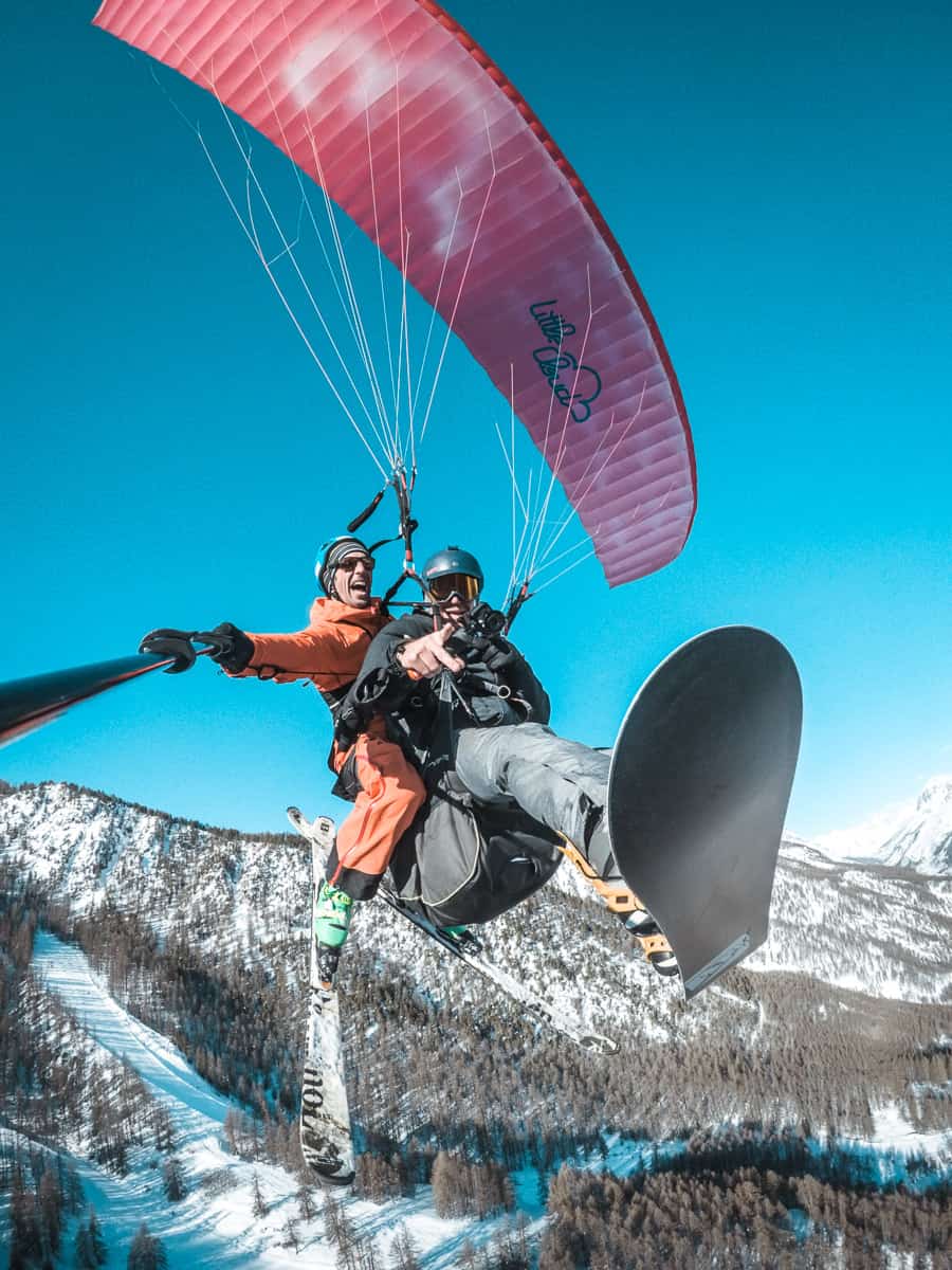 Jim fliegt mit Antoine von Axesse Parapente. Sie fliegen mit dem Gleitschirm über die Serre Chevalier Berge im Winter an einem Tag mit blauem Himmel.