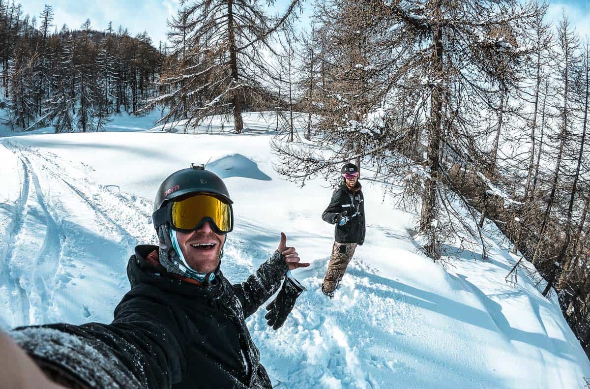 jim and loey are standing in fresh powder on a tree filled slope in serre chevalier. they're wearing their snowboarding gear. jim is smiling and doing a hangloose sign.