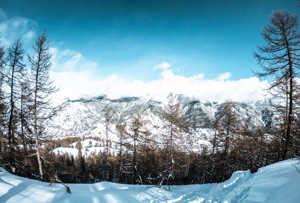 Die Berge von Serre Chevalier sind an einem sonnigen Tag mit Schnee bedeckt. Die Aufnahme entstand bei Jims Snowboardausflug abseits der Piste, daher ist im Vordergrund frischer Pulverschnee zu sehen und ein Wald, der den Hang hinunterführt.