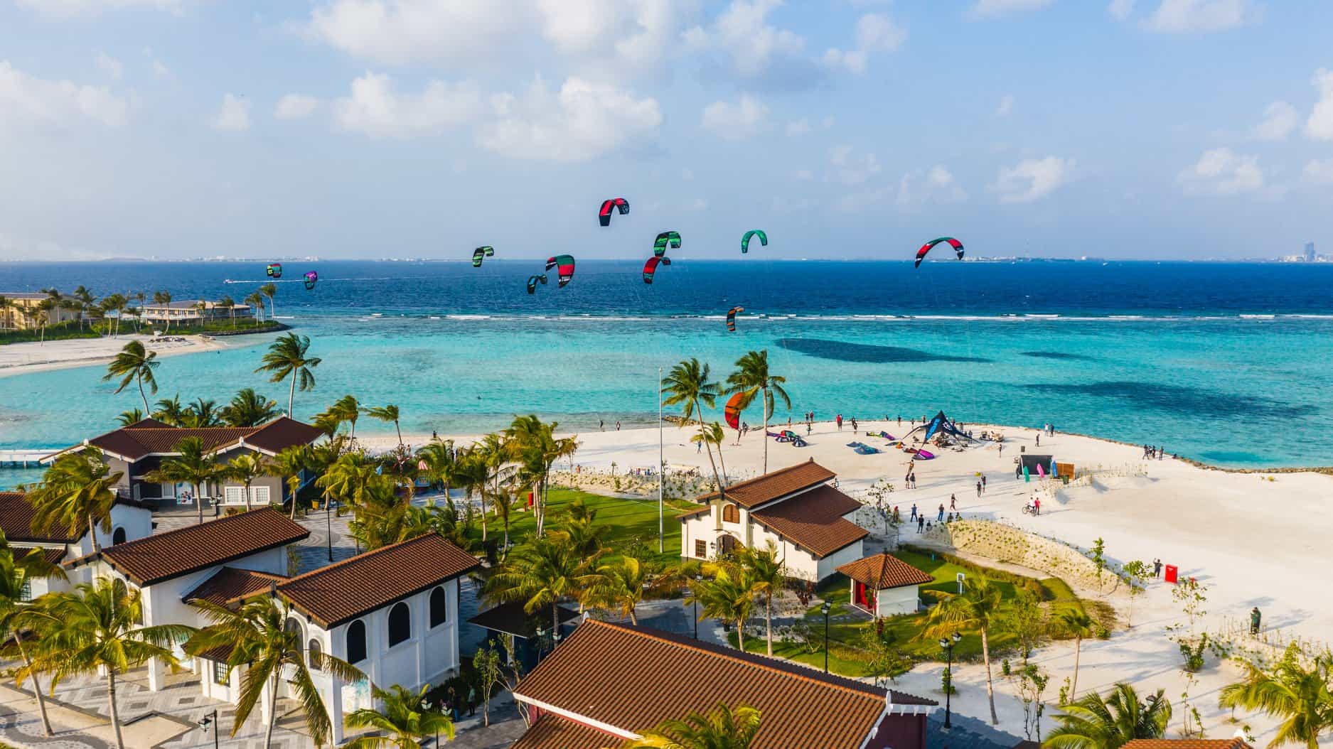 tropische kitesurfplek met een wit zandstrand, een paar luxe villa's aan de waterkant, en superblauw helder water. er vliegen een dozijn kites op het water.