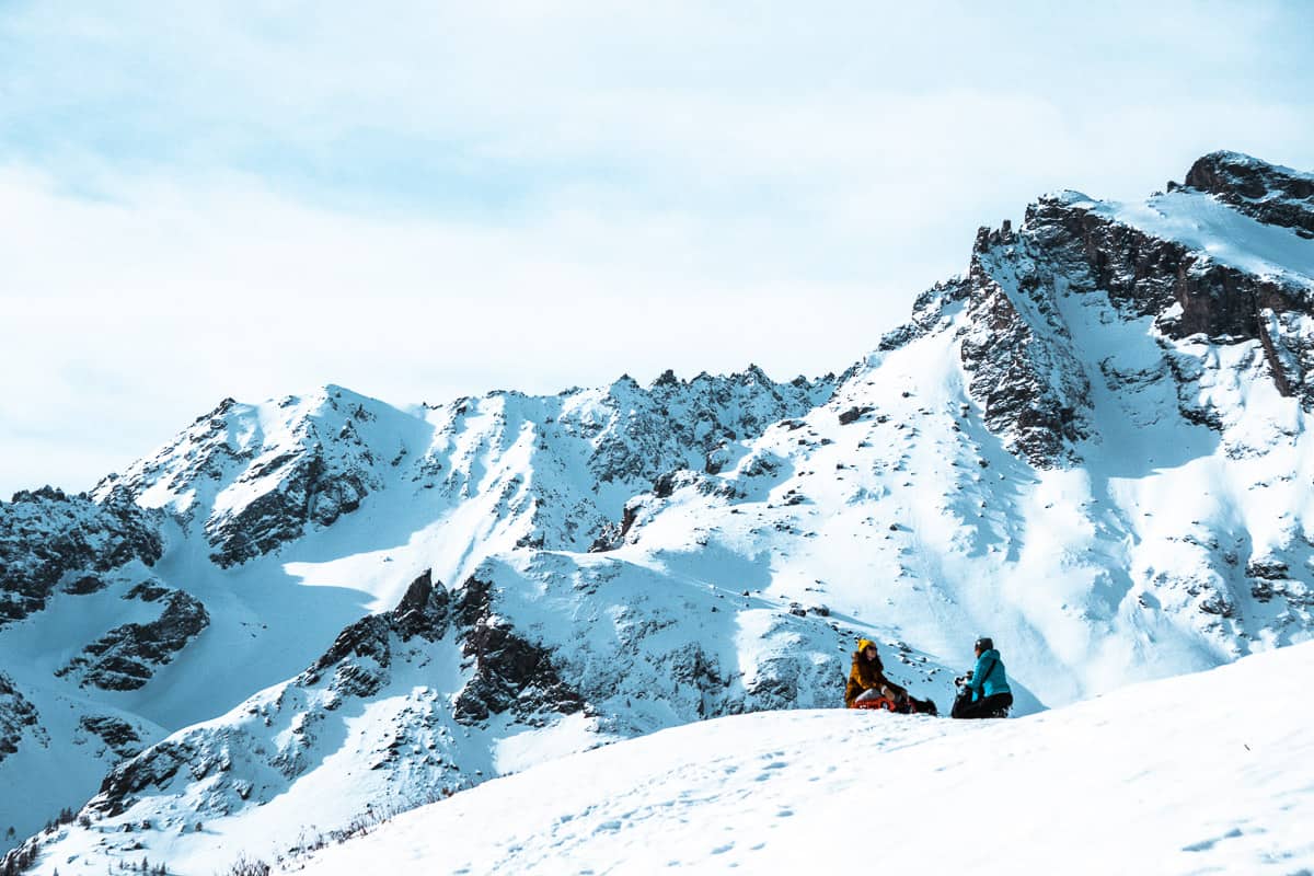 rachel and grace sitting on the snowy slopes of le monetier-les-bains preparing for meditation. the snow covered mountains behind look dramatic.