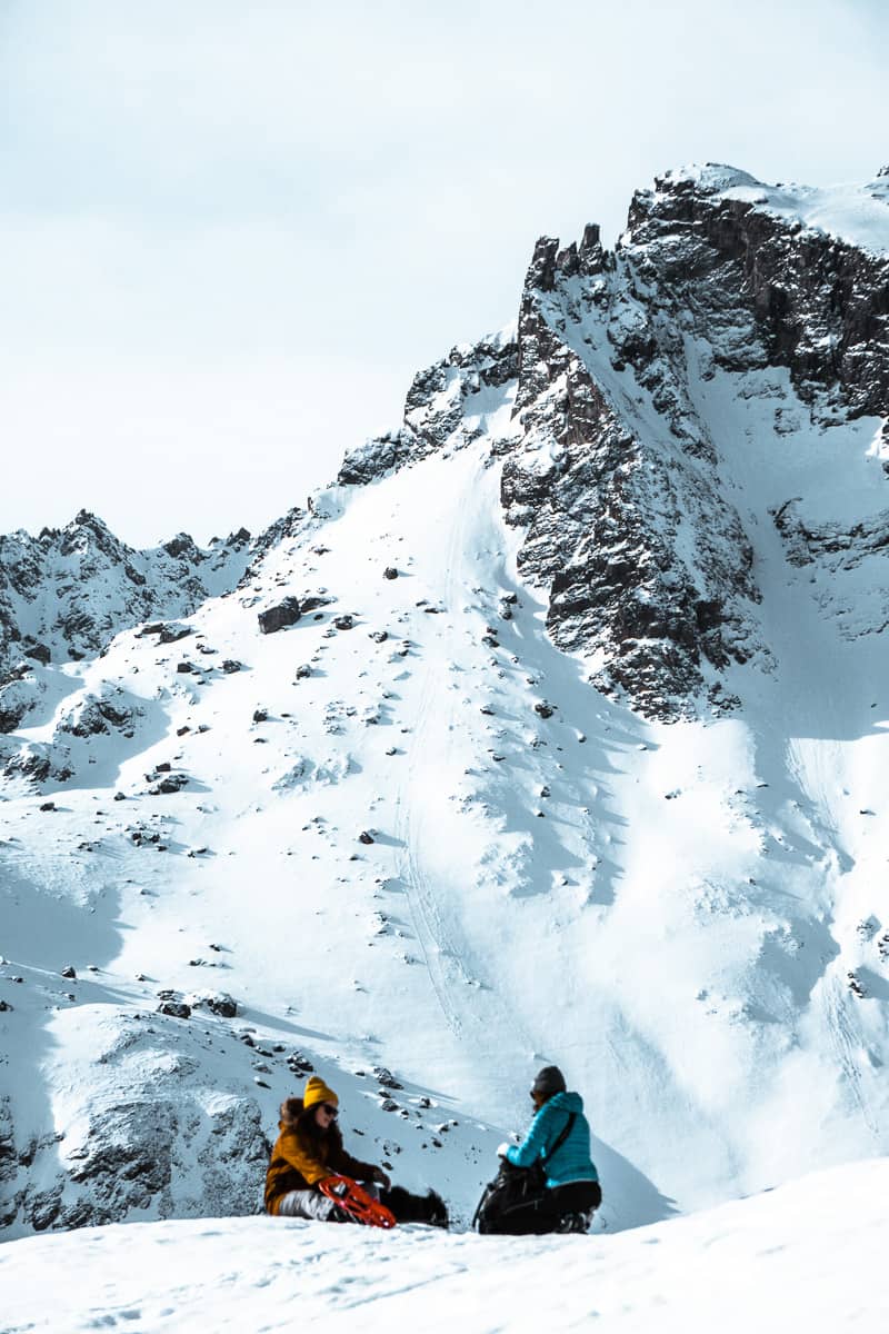 vertical shot of rachel and grace sitting in the snow at the foot of dramatic alpine mountains