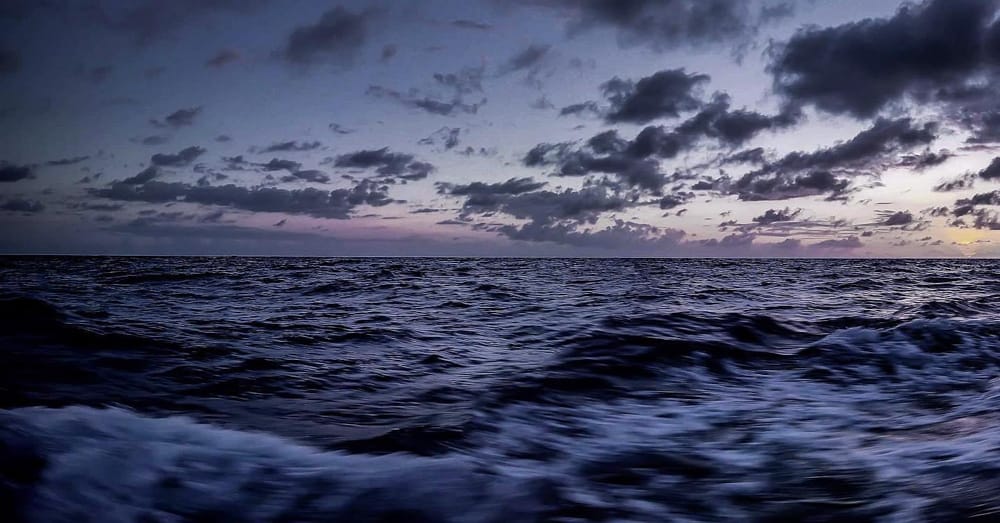 Die kabbelige Oberfläche des Ozeans bei Sonnenuntergang. Der Himmel und das Wasser haben eine tief violette Farbe, und der Himmel ist mit Wolkenfetzen gefüllt.