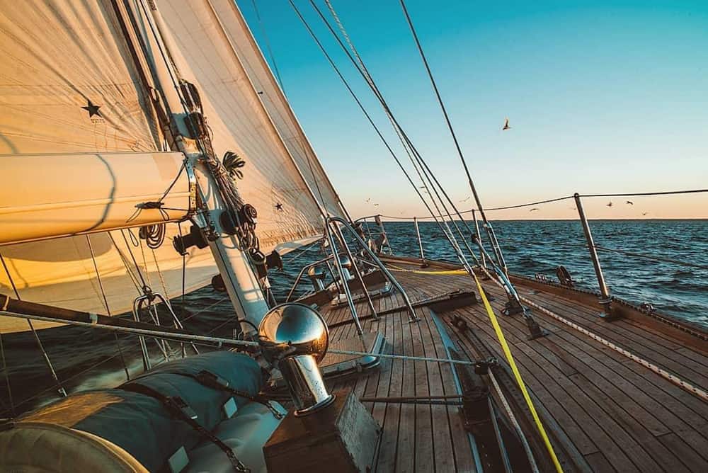 Opname genomen aan boord van het houten dek van een groot jacht. Het zeil is gehesen en meeuwen vliegen over het wateroppervlak net voorbij de boot.