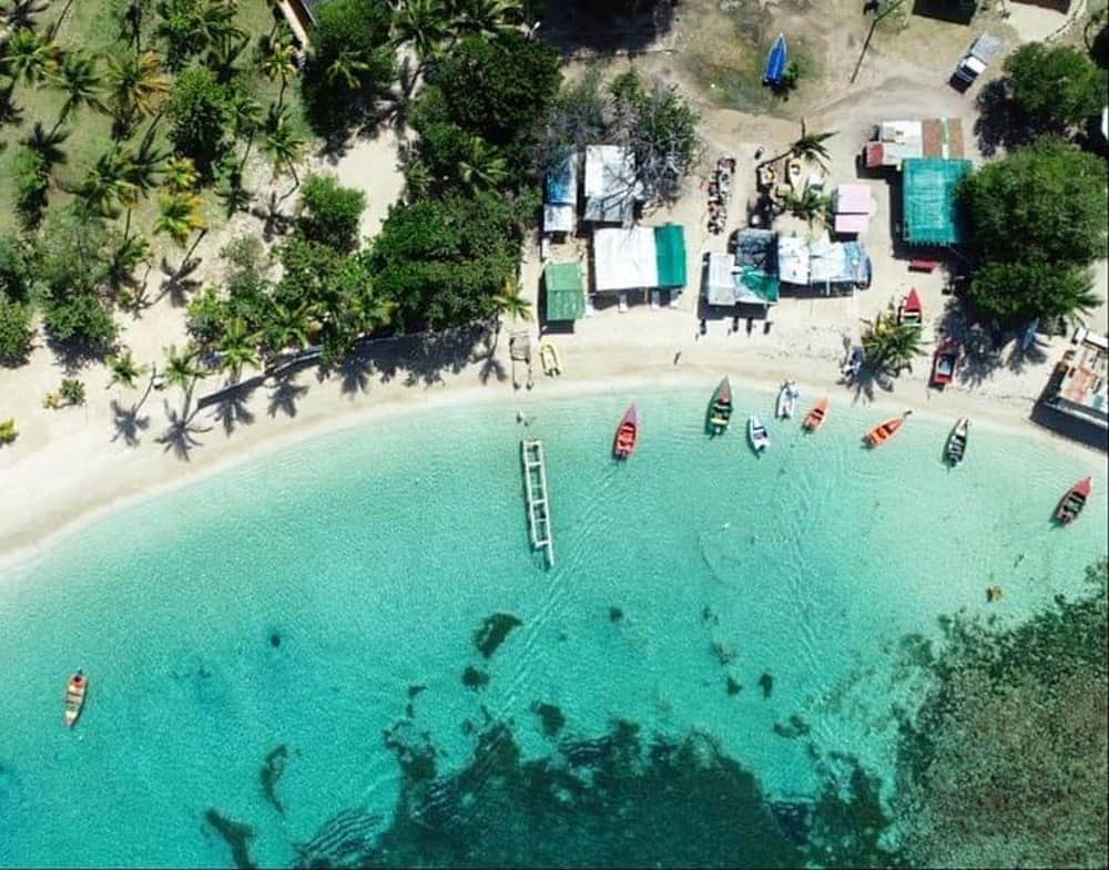 aerial shot of a tropical shoreline. There are some beach huts on the sand and a few colourful fishing boats moored just off the beach. There is a grove of palm trees on the left. The water is crystal clear and aqua coloured.