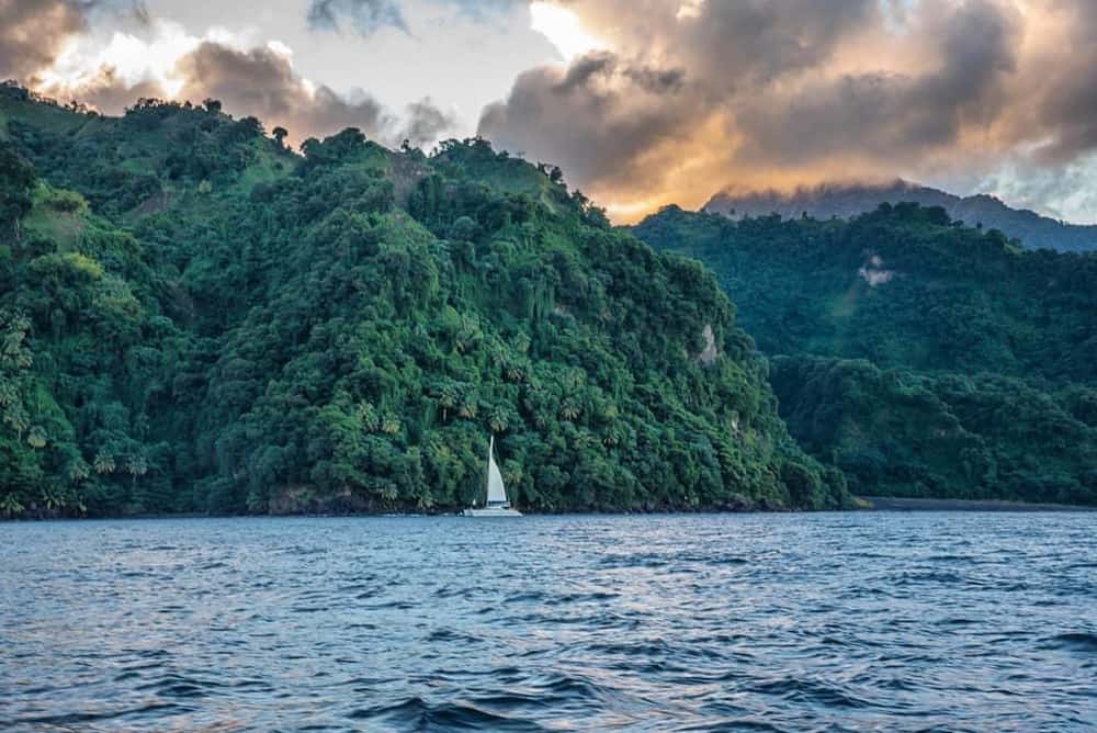 Eine weiße Yacht mit hochgezogenen Segeln, die vor der Küste eines tropischen, menschenleeren Gebiets vertäut ist. Der Himmel ist stimmungsvoll, es sieht aus wie die Zeit des Sonnenuntergangs. Das Wasser im Vordergrund trägt eine leichte Brandung.