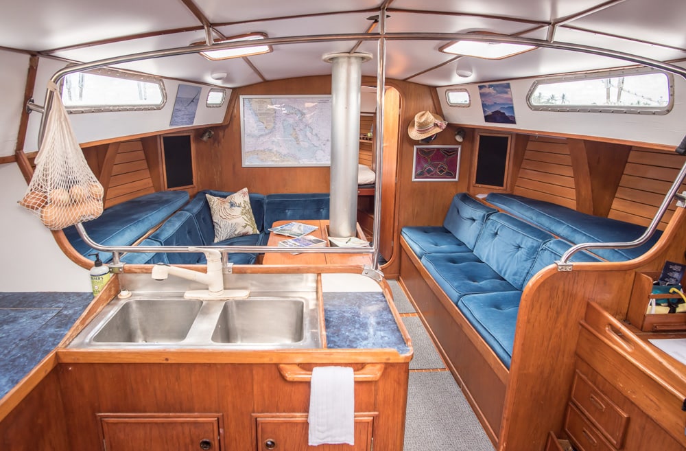 l'intérieur du bateau de Nathan et Vivian, Ultima. Il y a un évier en aluminium au premier plan à gauche, des canapés enveloppants en velours bleu au milieu de la photo, et la porte partiellement ouverte de ce qui semble être la chambre à coucher en arrière-plan.