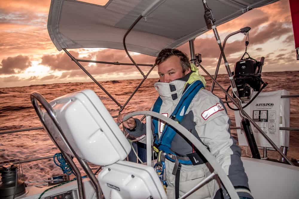 Nathan of Ocean passages suited up and on watch at the wheel of their boat. The sky and water behind him are lit up with a peachy pink sunset hue.