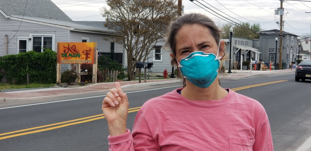 Rosa Linda wearing a pink tshirt and blue N95 face mask. She's standing on a road pointing at a Cinqo de Mayo sign behind her.