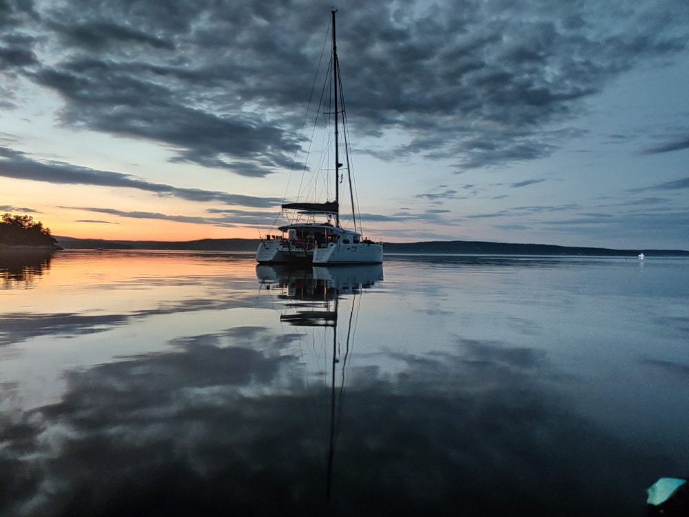 Rosa Lindas Katamaran liegt bei Sonnenuntergang auf spiegelglattem Wasser. Der Himmel ist in der Nähe des Horizonts schwach mit einem pfirsichfarbenen Farbton beleuchtet, und die Wolken spiegeln sich auf der ruhigen Wasseroberfläche.