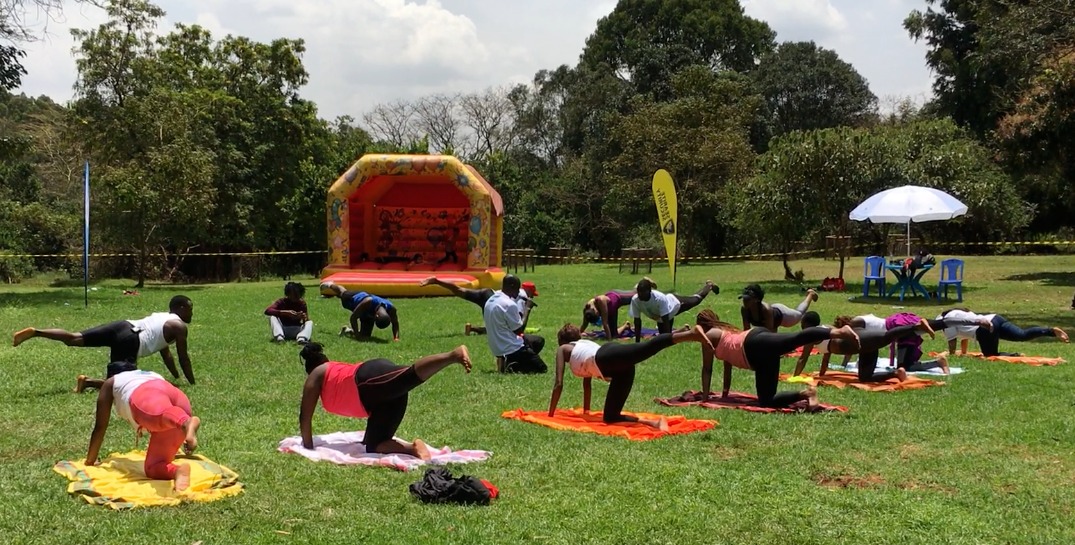 A woman dressed in black and white in a pool of light, in a seated yoga position with her palms raised upwards in front of her face. She's in a crowd of people doing yoga.